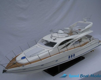 May include: White model yacht with a wooden deck and blue accents. The yacht has a large windshield and a small radar antenna on the top. The model is displayed on a black stand.