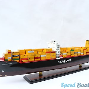 May include: A detailed model of a container ship, the "Hamburg Express", with a black hull and a red bow. The ship is loaded with yellow and red shipping containers and has the Hapag-Lloyd logo on the side. The model is displayed on a wooden base.