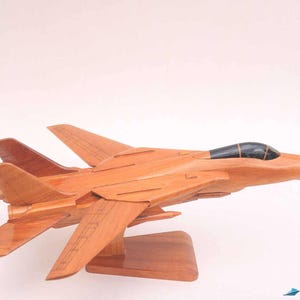 May include: A wooden model of a fighter jet, showcasing a detailed design with wings, tail fins, and a cockpit. The model is crafted from light brown wood and mounted on a wooden stand. The jet model is a collectible item.