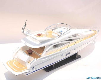 May include: White model yacht with a wooden deck and blue accents. The yacht has a large sun deck and a small table on the deck. The yacht is on a black stand.