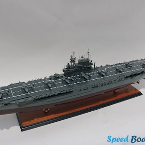 Könnte beinhalten: Ein detailliertes Modell eines grauen Flugzeugträgers mit einem Holzsockel. Der Träger hat ein Flugdeck mit Markierungen und mehreren kleinen Flugzeugen darauf.