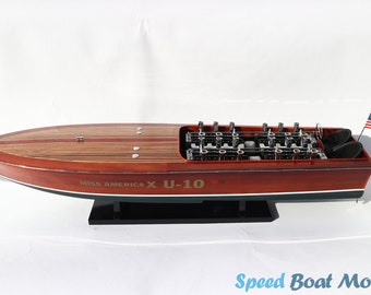 Può includere: Un modello in legno della barca veloce Miss America X U-10. Il modello è dipinto di rosso e nero con una striscia verde. La barca ha un motore dettagliato e una piccola bandiera americana sul retro.