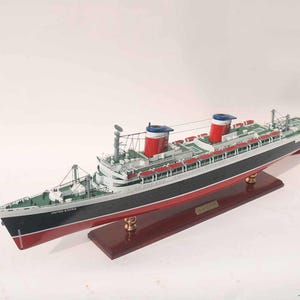 May include: A detailed model of the United States ocean liner. The ship features a black hull with red accents, a white superstructure, and red and white smokestacks with blue tops. The model sits on a wooden base and includes the ship's name.