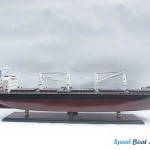 Pode incluir: Um modelo detalhado de um grande navio de carga com uma superestrutura branca e um casco vermelho escuro. O navio possui várias gruas e é apresentado em uma base de madeira.