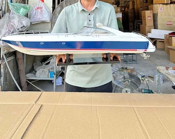 May include: A scale model motor yacht featuring a white upper deck and a deep blue hull with a thin red stripe along the waterline. The vessel includes a clear windshield, silver railings, and a black display stand, showcasing a sleek design.