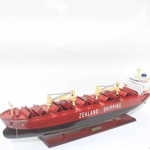 Pode incluir: Um modelo de navio vermelho e preto com o texto "ZEALAND SHIPPING" na lateral. O modelo é montado em uma base de madeira. O navio tem guindastes amarelos e vários contêineres de carga.