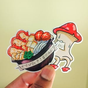 May include: A sticker featuring a cartoon mushroom with a red cap and a bowl of Japanese hot pot with the text "ナハハと寿の火は" (Nahaha to Kotobuki no Hi wa) which translates to "Laughter and the fire of longevity". The mushroom has a note that says "맛있어" (Masisseo) which translates to "Delicious" in Korean.