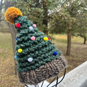 May include: A handmade Christmas tree hat with a brown knitted brim and a green tree-shaped crown. The hat is adorned with colorful pom-poms and a yellow pom-pom at the top. The hat is displayed on a black metal stand.
