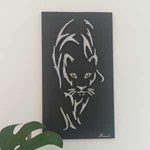 Könnte beinhalten: Ein schwarzes, rechteckiges Wandkunstwerk mit einer stilisierten silbernen Umrisszeichnung eines Panthers. Das Kunstwerk ist an einer weißen Wand angebracht, mit einer großen grünen Pflanze im Vordergrund. Die Signatur des Künstlers befindet sich in der unteren rechten Ecke.