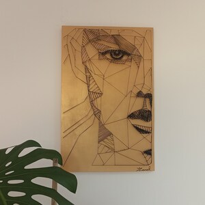 Könnte beinhalten: Ein geometrisches Porträt eines Gesichts ist auf einer goldfarbenen rechteckigen Tafel dargestellt. Das Kunstwerk zeigt ein Halbgesichtsdesign mit kantigen Linien und Schattierungen, wodurch eine moderne Ästhetik entsteht. Das Stück ist unten rechts signiert.