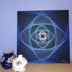 Könnte beinhalten: Eine quadratische, schwarze Leinwand mit einem geometrischen String-Art-Design in Blautönen. Das Kunstwerk zeigt ein zentrales Quadrat mit radialen Linien, die eine faszinierende optische Täuschung erzeugen. Zwei dekorative Keramikteile befinden sich im Vordergrund.