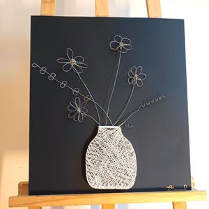 Könnte beinhalten: Eine weiße String-Art-Vase vor schwarzem Hintergrund mit silbernen Drahtblumen. Die Vase steht auf einem Holzstaffelei.