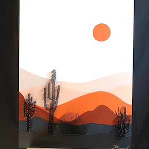 Könnte beinhalten: Eine Wüstenlandschaft mit einer roten Sonne, orangefarbenen und braunen Hügeln und schwarzen Drahtkakteen. Das Kunstwerk ist auf einer weißen Leinwand mit einem schwarzen Rahmen.