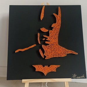 Könnte beinhalten: Batman-String-Art auf schwarzem Hintergrund. Das Kunstwerk zeigt eine orangefarbene Fadenkontur von Batmans Gesicht und dem Fledermaussymbol. Präsentiert auf einer kleinen Holzstaffelei.