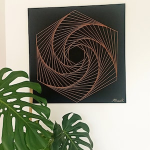 Könnte beinhalten: Geometrische Wandkunst mit einem spiralförmigen Design aus kupferfarbenen Linien auf einer schwarzen quadratischen Leinwand. Das Kunstwerk hängt an einer weißen Wand, mit großen grünen Monstera-Blättern im Vordergrund.