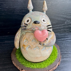 Può includere: Statuetta di Totoro in ceramica con un cuore rosa. La figura è grigio chiaro con dettagli beige e marroni, baffi neri e grandi occhi rotondi. Poggia su una base di legno con muschio verde.