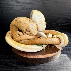 May include: A ceramic sculpture of a coiled snake with a light brown, speckled body and a cream-coloured head. The snake has a golden eye and rests on a wooden base. The sculpture is a decorative art piece.