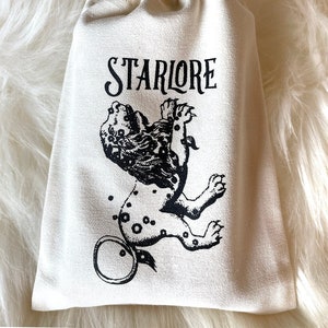 May include: A white canvas drawstring bag with the text "STARLORE" printed in black. A black and white illustration of a lion with a ring around its tail is printed on the bag.