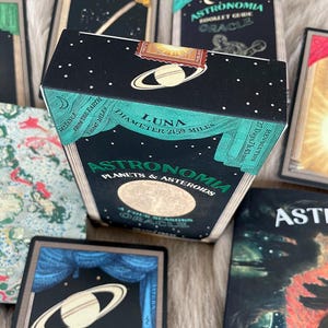 May include: A black box with gold and green accents featuring the text "Astronomia Planets & Asteroids" and "Luna Diameter 2159 Miles". The box is decorated with a celestial scene and stars.