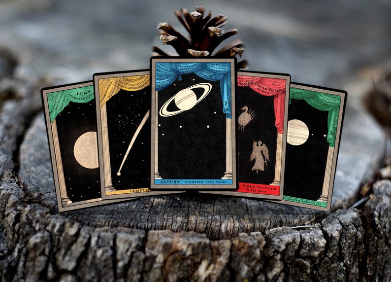 May include: Five vintage playing cards with celestial imagery. Each card features a different celestial body, including the moon, a comet, Saturn, a constellation, and a star. The cards are decorated with ornate frames and have a dark background.