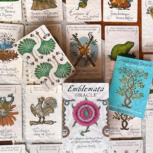 May include: A collection of Emblemata Oracle cards featuring Renaissance-style illustrations, including a central card with green peacock feather fans, a deck box with a pink rose emblem, and a blue keywords guide booklet with a tree illustration.