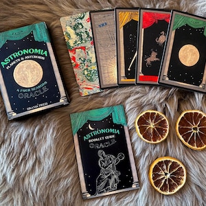 May include: A green and black box with the title "Astronomia Planets & Asteroids A Four Seasons Oracle" and the text "Urania Press". The box is surrounded by five cards with black backgrounds and gold accents. The cards feature celestial imagery, including planets, stars, and moons. The cards are arranged in a semi-circle around the box.
