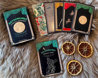 Może przedstawiać: Zielone i czarne pudełko z tytułem "Astronomia Planets & Asteroids A Four Seasons Oracle" i tekstem "Urania Press". Pudełko jest otoczone pięcioma kartami z czarnymi tłami i złotymi akcentami. Karty przedstawiają obrazy niebieskie, w tym planety, gwiazdy i księżyce. Karty są ułożone w półkole wokół pudełka.