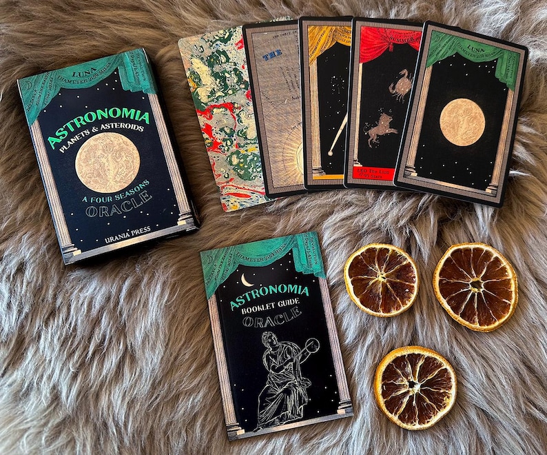 May include: A green and black box with the title "Astronomia Planets & Asteroids A Four Seasons Oracle" and the text "Urania Press". The box is surrounded by five cards with black backgrounds and gold accents. The cards feature celestial imagery, including planets, stars, and moons. The cards are arranged in a semi-circle around the box.