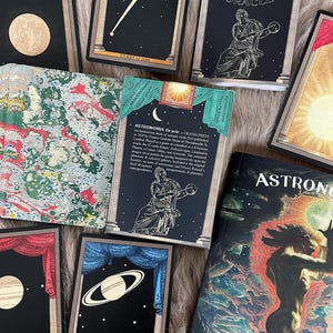 May include: A set of black and gold tarot cards with illustrations of planets and celestial bodies. The cards are part of an oracle deck called "Astronomia Oracle" and feature a booklet guide. The cards are laid out on a fuzzy brown surface.