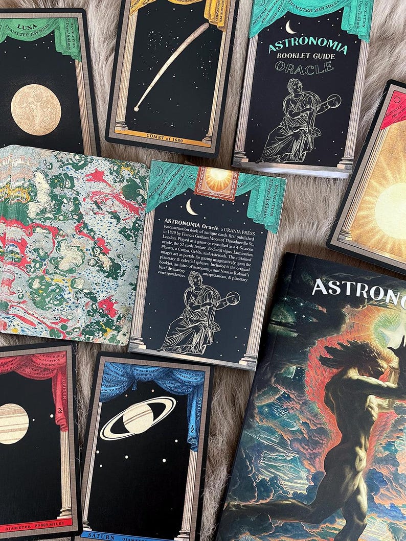 May include: A set of black and gold tarot cards with illustrations of planets and celestial bodies. The cards are part of an oracle deck called "Astronomia Oracle" and feature a booklet guide. The cards are laid out on a fuzzy brown surface.