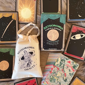 May include: An oracle deck with astronomy-themed cards and a drawstring bag. The cards feature celestial bodies and constellations, with titles like "The Sun" and "Venus." The bag is cream-colored and has the words "Astronomia Oracle" printed on it.