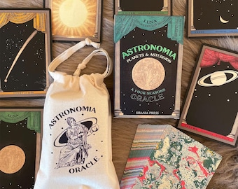 May include: An oracle deck with astronomy-themed cards and a drawstring bag. The cards feature celestial bodies and constellations, with titles like "The Sun" and "Venus." The bag is cream-colored and has the words "Astronomia Oracle" printed on it.