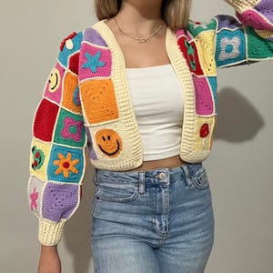 Könnte beinhalten: Eine bunte gehäkelte Strickjacke mit Patchwork-Design mit Smiley-Gesichtern, Blumen und Sternen. Die Strickjacke ist kurz und hat lange Ärmel.