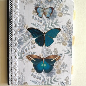 May include: A decorative journal with a white cover featuring three butterfly illustrations in shades of blue and brown. The cover has a floral pattern and is accented with a white lace trim along the spine. The journal has three beige tabs.