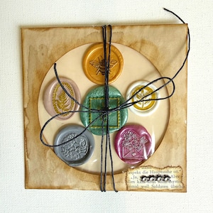 May include: A set of six wax seals in various colors, including gold, green, pink, and gray, each with a different embossed design. The seals are arranged in a circle within a tan paper frame, tied with black string. A piece of paper with text is visible.