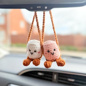 May include: Two crocheted car charms, one white and one pink, each with smiling faces and brown legs. They hang from the rearview mirror by brown cords. The car interior is visible in the background.