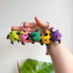 May include: Five colorful crochet keychains in green, pink, green, yellow, and purple. Each keychain features a small, round creature with black legs, arms, and eyes. The keychains are held by a person's hand.