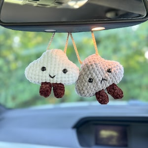 May include: Two crocheted cloud car charms suspended from a car's rearview mirror. One is white with a smiling face and brown feet, the other is grey with a sad face and brown feet. They hang by beige cords.