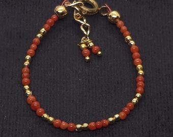 Puede incluir: Un brazalete delicado hecho de pequeñas cuentas de coral rojo y cuentas doradas. El brazalete tiene un cierre dorado y un pequeño dije con tres cuentas de coral.