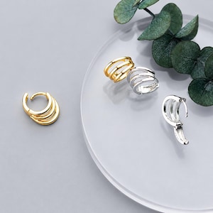 May include: Three hoop earrings featuring a triple-band design. One earring is gold-toned and positioned separately, while two earrings are silver-toned, with one shown closed and the other partially open, revealing a hinged mechanism.