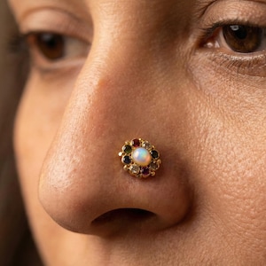Puede incluir: Primer plano de un piercing de nariz dorado con una piedra central de ópalo rodeada por un halo de piedras preciosas de colores. El piercing está colocado sobre la piel de la nariz de una persona, con el ojo y la piel circundante visibles en el fondo.