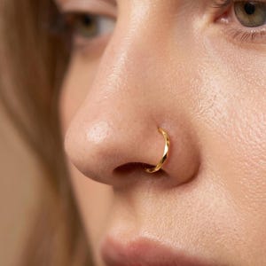 May include: Close-up of a nose with a gold-colored nose ring. The ring is a small, curved hoop with a textured, faceted design. The skin around the nose is smooth and fair, with a slight shadow.