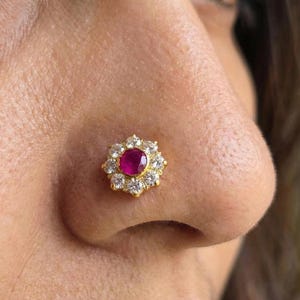May include: A gold nose stud featuring a central, round, deep pink gemstone surrounded by a halo of smaller, clear gemstones. The stud is set against the skin of a person's nose, showcasing the jewelry's design and sparkle.