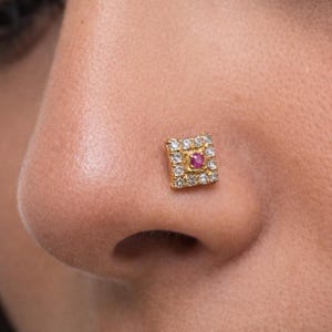 May include: A square gold nose stud with a central red gemstone, surrounded by smaller clear stones. The jewelry is set against the skin of a person's nose, showcasing the stud's intricate design and sparkle.