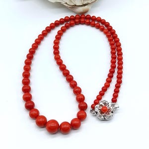 Può includere: Una collana composta da una fila di perle rotonde rosse graduate. Le perle aumentano di dimensioni verso il centro della fila. La collana è chiusa da una chiusura in metallo argentato a forma di fiore con una piccola perla rossa al centro.