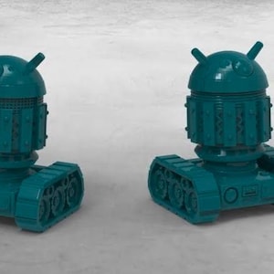 May include: Two teal robot figurines, each with a rounded head and antenna-like protrusions, are positioned on a light grey surface. The robots have a cylindrical midsection and track-like treads, resembling a tank. The design is a playful, stylized representation.