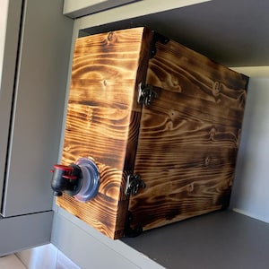 Op de afbeelding: Een houten doos met een gebrande afwerking en metalen scharnieren. De doos heeft een tuit aan de zijkant om vloeistoffen te schenken.