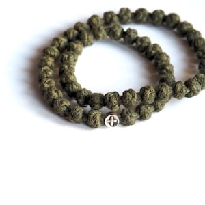 Può includere: Un bracciale circolare realizzato in corda annodata color verde oliva, caratterizzato da un piccolo ciondolo rotondo in metallo argentato con un simbolo a croce in rilievo sulla superficie. La corda è testurizzata e formata da nodi.