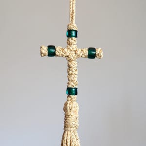 May include: A beige macrame cross with dark green glass beads. The cross is suspended from a matching cord, featuring a knotted design. The cross is a decorative item.