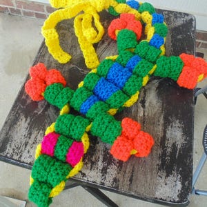 May include: A colorful, handmade crochet lizard toy. The lizard features a green body with blue and yellow accents, orange feet, and a yellow bow. The toy is placed on a weathered, dark-colored table.